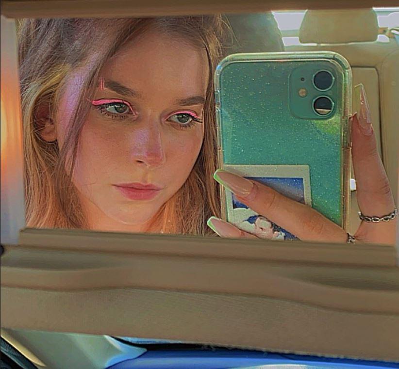 The second Hannah Petersen takes a selfie in a car mirror.  She wore neon pink eyeliner on both her eyes and brows.