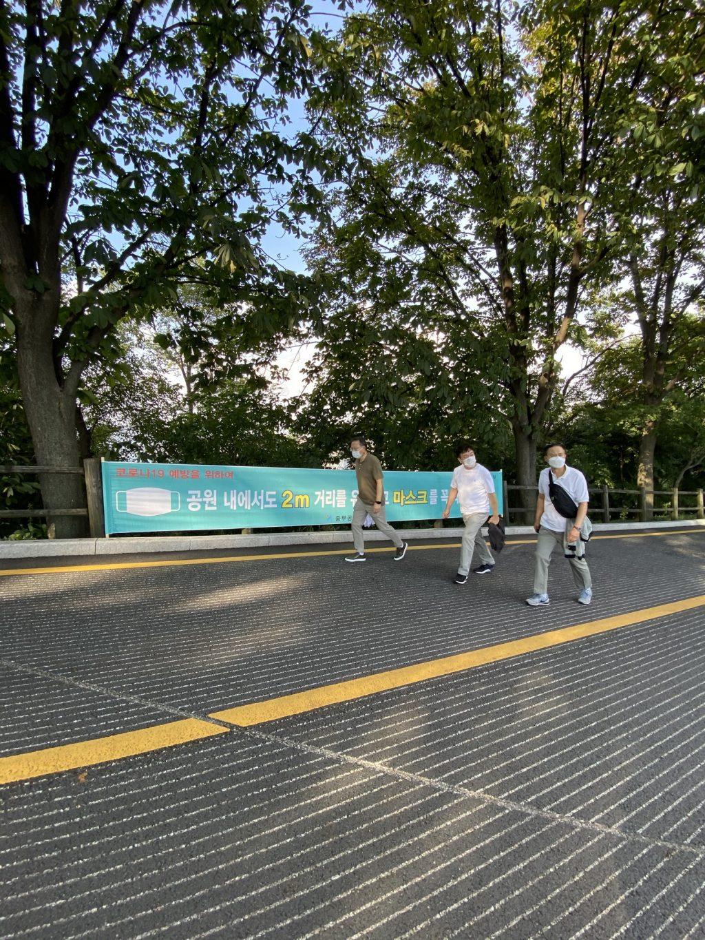 Three men wear masks while walking in front of a banner that reminds the public of the COVID-19 precautions. Similar forms of this banner were placed along both sides of the road with occasional hand sanitizers on small stools.