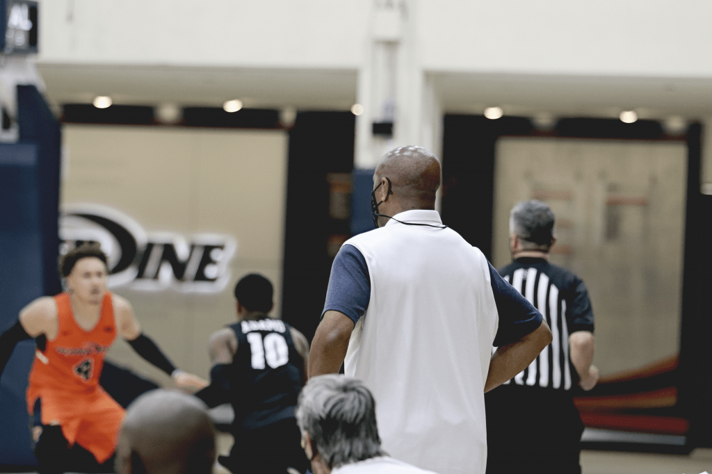 Head Coach Lorenzo Romar observes the Waves team from the sideline, while senior guard Colbey Ross defends Portland guard XXX.