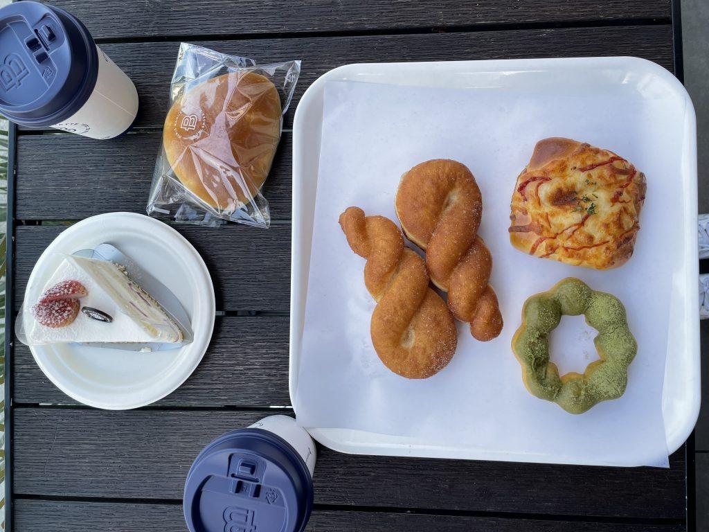 A slice of strawberry soft cream cake, chocolate cream bread, a matcha mochi donut, two twisted donuts, a hashbrown ham and cheese bread and a vanilla latte rest on the table before being consumed. These foods were my favorites growing up.