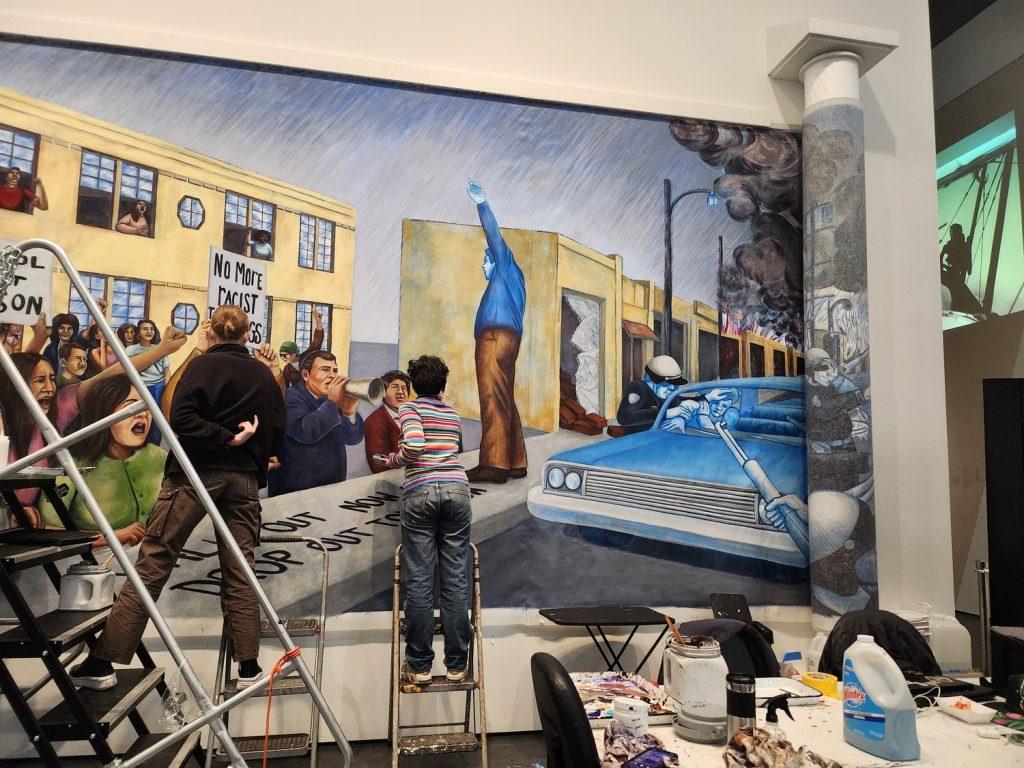 Artists Lovelace (left) and Kira Layton paint a portion of the mural portraying the 1968 East L.A. Walkouts melting into the Watts Rebellion on March 29, in LACMA. The rolled-up canvas to the right of the surface the artists are currently working on allows them to work on an area much larger than the wall of the museum, Mendoza said.